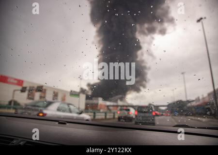 À travers un pare-brise moucheté par la pluie, une colonne massive de fumée noire s'élève d'un incendie industriel au loin, provoquant des embouteillages en voiture Banque D'Images