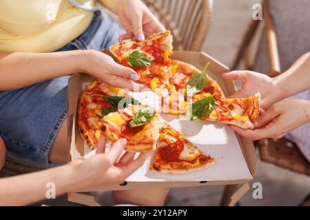 Un groupe d'amis partagent la pizza à l'extérieur, les mains cherchant des tranches garnies de basilic frais, tandis qu'une boîte à pizza et des assiettes de hors-d'œuvre s'assoient à l'arrière Banque D'Images