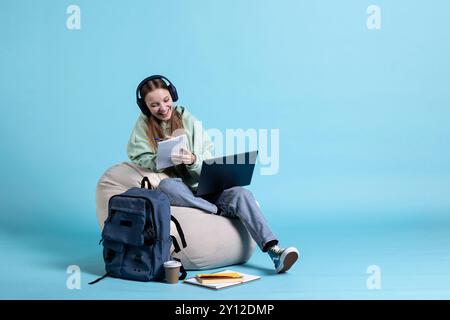 Fille souriante utilisant un ordinateur portable pour apprendre des informations pour les examens du lycée tout en écoutant de la musique, fond de studio. Élève adolescent optimiste entendant de la musique et tenant un cahier, écrivant sur le bloc-notes Banque D'Images