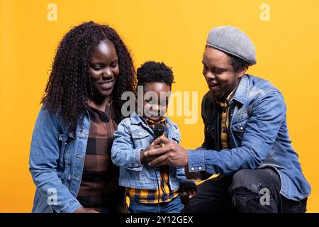 Fiers parents écoutant leur fils bébé chanter une chanson au micro, riant et étant adorables ensemble sur fond jaune. Heureuse famille afro-américaine de trois aiment la musique. Banque D'Images