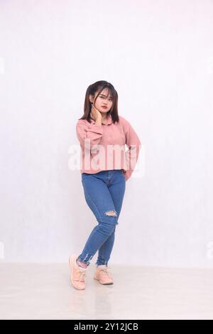 Souriant gai aux cheveux noirs belle femme dans ses années 20 portant une chemise rose de base debout style de travail tout en tenant le cou et les hanches fond blanc Banque D'Images