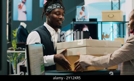 Employé de vente au détail ramassant les boîtes de commande de vêtements dans le grand magasin, signant la facture et les papiers de confirmation à la caisse enregistreuse. Femme afro-américaine donnant la marchandise demandée. Caméra A. Banque D'Images