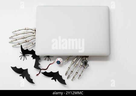 Composition avec ordinateur portable moderne, mains squelettes et décorations d'Halloween sur fond blanc Banque D'Images