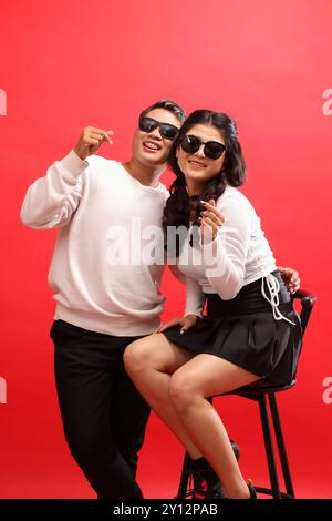 Une paire de jeunes amants, un joyeux homme asiatique et une femme portant des chandails blancs, des lunettes de soleil, la femme assise sur une chaise à côté de l'homme tout en montrant Banque D'Images