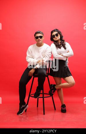 Corps complet d'une paire de jeunes amoureux asiatiques élégants portant des chandails blancs, des lunettes de soleil, l'homme assis dans une chaise tandis que la femme est penchée en arrière. pour Banque D'Images