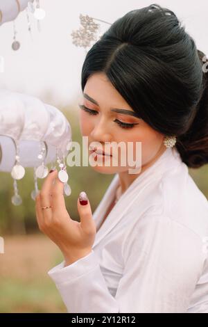 Charmante jeune femme asiatique debout regardant et tenant un parapluie portant des vêtements traditionnels javanais modernes, kebaya de velours blanc marié dans un out Banque D'Images