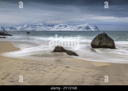 Rochers sur la plage en face de montagnes enneigées, nuages sombres, mer, jet, hiver, Vikten, Flakstadoya, Lofoten, Norvège, Europe Banque D'Images