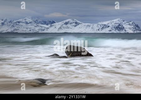 Rochers sur la plage en face de montagnes enneigées, nuages sombres, mer, jet, hiver, Vikten, Flakstadoya, Lofoten, Norvège, Europe Banque D'Images