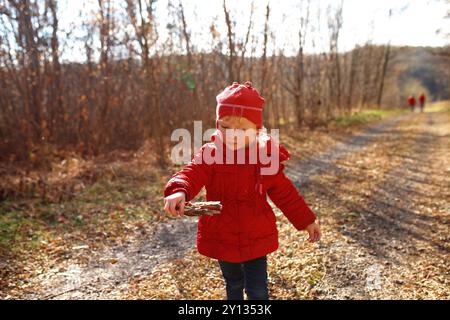 Un enfant vêtu d'un manteau rouge vif marche le long d'un chemin forestier, rassemblant des bâtons au milieu d'un feuillage d'automne coloré sous un ciel clair. Banque D'Images