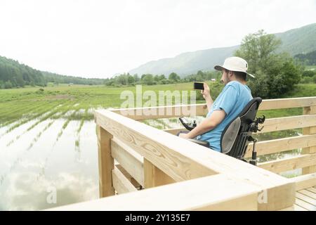 Homme en fauteuil roulant en utilisant la caméra de smartphone près du lac dans la nature Banque D'Images