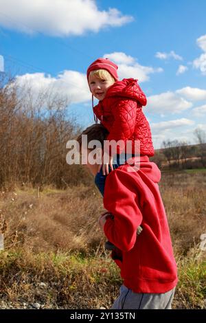 Un jeune garçon marche avec sa petite sœur sur ses épaules, partageant un moment de joie au milieu d’un beau paysage automnal sous un ciel bleu. Banque D'Images