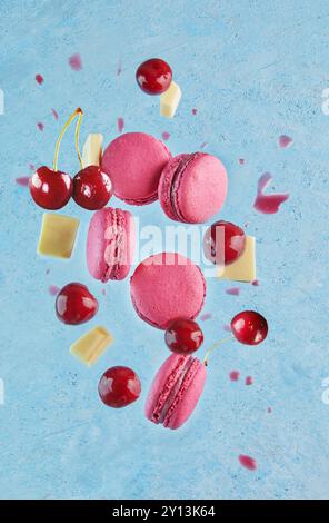 Composition artistique en lévitation de macarons roses, de cerises fraîches et de morceaux de chocolat blanc volant dans les airs sur fond bleu. Banque D'Images