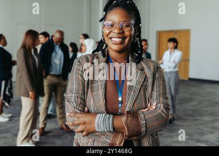 Femme noire confiante avec des cheveux tressés et des lunettes debout à un événement de réseautage de conférence d'affaires. Groupe diversifié de professionnels dans le backgrou Banque D'Images