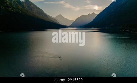 Un paddleboarder solitaire glisse sur les eaux calmes de Plansee au lever du soleil, tandis que le soleil jette une lueur chaude sur le lac et met en valeur les superbes montagnes entourant cette oasis de sérénité. Banque D'Images