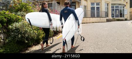 Une image panoramique de deux surfeurs portant leurs planches de surf marchant vers le haut de GT Great Western Beach à Newquay en Cornouailles au Royaume-Uni. Banque D'Images