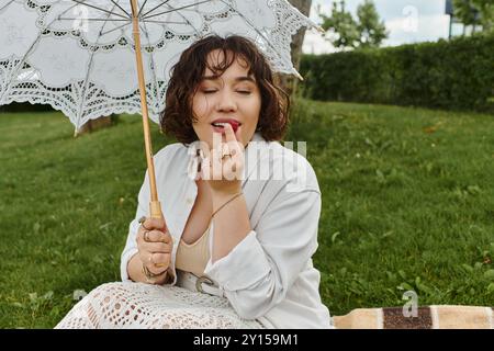 Une charmante jeune femme profite d'un moment serein, en appliquant un baume à lèvres sous son délicat parasol au soleil d'été. Banque D'Images