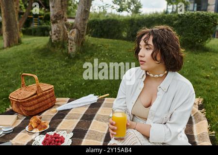 Une jeune femme profite d'un moment de paix, sirotant du jus tout en se reposant sur une couverture de pique-nique entourée par la nature. Banque D'Images