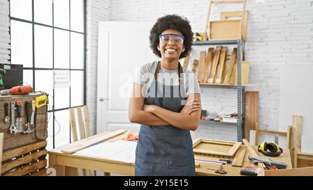 Une femme africaine confiante portant des lunettes de sécurité et un tablier en denim se tient les bras croisés dans un atelier de menuiserie bien éclairé. Banque D'Images