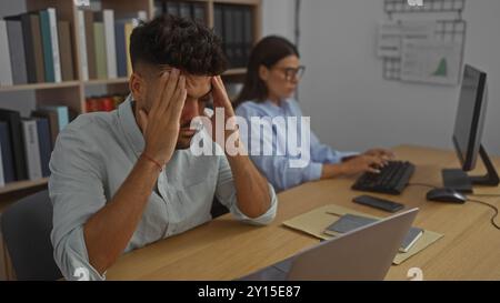 Homme stressé à son bureau tandis que la femme travaille en arrière-plan dans un bureau, les deux se concentrant sur les tâches informatiques au milieu d'une charge de travail occupée Banque D'Images