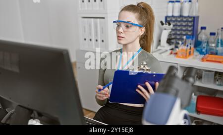 Une jeune femme concentrée avec des lunettes de sécurité et une planchette à pince mène des recherches dans un laboratoire bien équipé. Banque D'Images