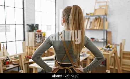 Vue arrière d'une jeune femme blonde debout avec confiance dans un atelier de menuiserie lumineux, les mains sur les hanches. Banque D'Images