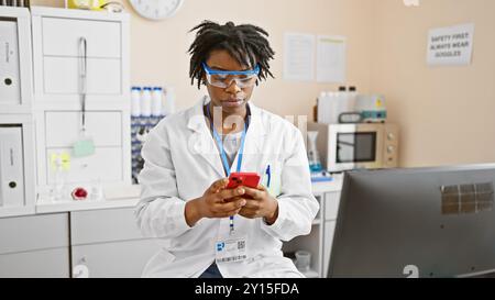 Afro-américaine scientifique avec des dreadlocks portant des lunettes de sécurité et blouse de laboratoire à l'aide d'un smartphone en laboratoire. Banque D'Images