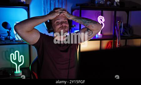 Homme tatoué avec des écouteurs dans une salle de jeu éclairée au néon l'air frustré ou fatigué tout en tenant sa tête. Banque D'Images