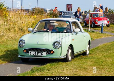 Une Nissan Figaro vert clair classique conduite par une journée ensoleillée avec des gens et une autre voiture vintage en arrière-plan. Banque D'Images