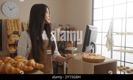 Jeune femme chinoise travaillant dans une boulangerie, debout près du comptoir à l'intérieur avec divers étalages de pain autour d'elle et un intérieur ensoleillé avec une horloge allumée Banque D'Images