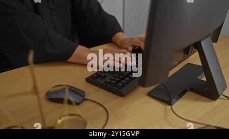 Juge mains tapant sur un clavier dans un cadre de bureau, avec des échelles de justice sur le bureau en bois, mettant l'accent sur l'environnement de travail judiciaire Banque D'Images