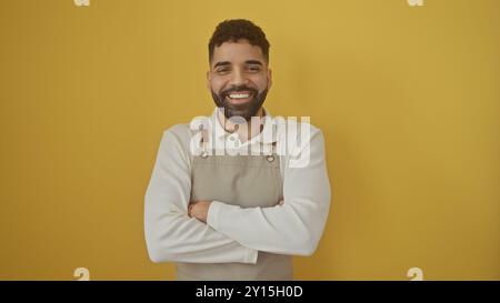 Un jeune homme hispanique joyeux avec une barbe portant un tablier se tient sur un fond jaune isolé, les bras croisés, dégageant une ambiance amicale. Banque D'Images