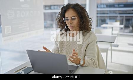 Femme hispanique d'âge moyen qui passe un appel vidéo depuis un bureau moderne, illustrant ainsi la communication sur le lieu de travail. Banque D'Images