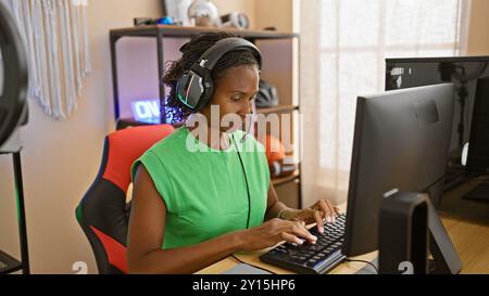 Femme afro-américaine portant des écouteurs travaillant intensément dans un bureau à domicile moderne avec configuration de jeu. Banque D'Images