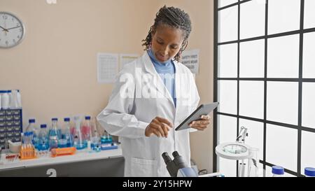 Une scientifique afro-américaine analyse les données sur une tablette dans un laboratoire, entourée d'équipements et d'échantillons. Banque D'Images