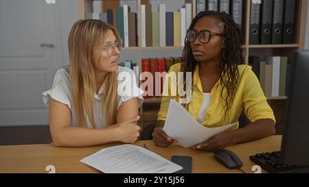 Femmes discutant des documents d'affaires à une table de bureau avec des livres et des dossiers en arrière-plan Banque D'Images