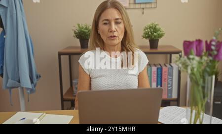 Femme blonde mature travaillant sur ordinateur portable dans un bureau à domicile avec des cahiers et des fleurs, exsudant concentration et tranquillité Banque D'Images