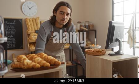 Bel homme dans un tablier travaillant dans une boulangerie avec des étagères de pain et de pâtisseries, debout à l'intérieur à côté d'une caisse enregistreuse Banque D'Images