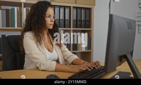 Jeune femme aux cheveux bruns bouclés travaillant à un bureau dans un cadre de bureau avec des étagères derrière elle Banque D'Images