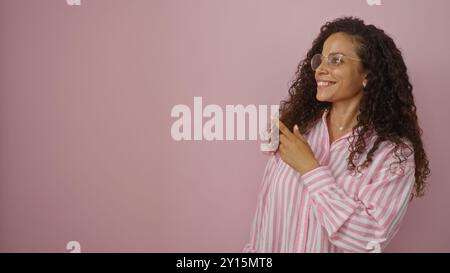 Jeune femme hispanique attrayante avec des pointes de cheveux bouclés sur le côté tout en souriant sur un fond rose isolé Banque D'Images