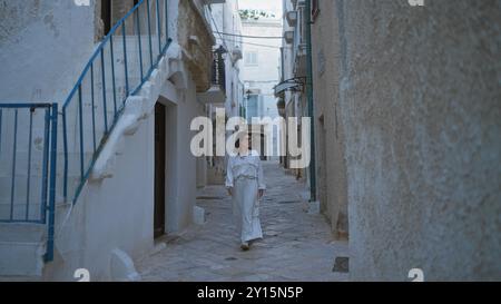 Une jeune et belle femme hispanique se promène dans les charmantes rues pavées de pierre de polignano a mare, pouilles, italie, habillée de blanc et entourée de b. Banque D'Images