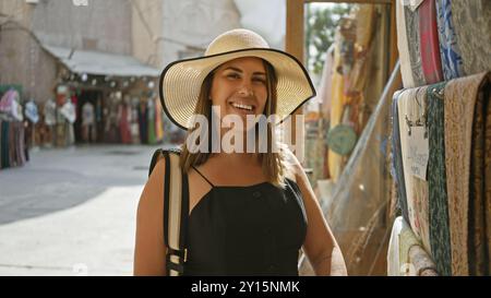 Une jeune femme souriante avec un chapeau de soleil explore un souk traditionnel de dubaï, entouré de textiles vibrants. Banque D'Images