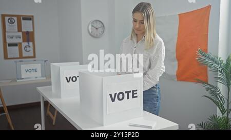 Une jeune femme blonde et séduisante vote dans un collège électoral en intérieur en irlande, avec un drapeau irlandais et une horloge en arrière-plan. Banque D'Images