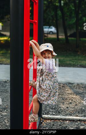 Jeune fille dans une robe florale et chapeau de soleil grimpant une structure d'échelle rouge à une aire de jeux en plein air sur une journée ensoleillée. Banque D'Images