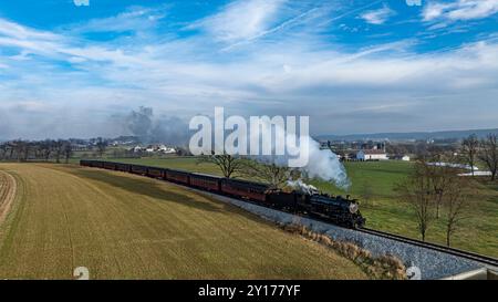 Une locomotive à vapeur d'époque longe des voies ferrées pittoresques, dégageant de la fumée blanche lorsqu'elle traverse des champs verts. Le paysage présente des arbres lointains et un ciel bleu clair. Banque D'Images