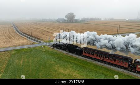 Un train à vapeur vintage souffle d'épais nuages de fumée alors qu'il chute à travers un cadre rural brumeux, entouré de vastes champs et de structures agricoles lointaines sous un ciel gris. Banque D'Images