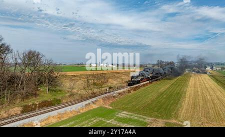 Une locomotive à vapeur vintage descend le long de la voie ferrée entourée de vastes champs verts et de fermes par une journée claire et lumineuse, émettant des panaches de fumée grise dans le ciel. Banque D'Images