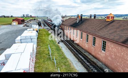 Un train à vapeur vintage souffle de la fumée blanche alors qu'il se déplace le long des voies à côté d'un bâtiment en briques rouges dans une zone rurale pittoresque sous un ciel nuageux. Banque D'Images