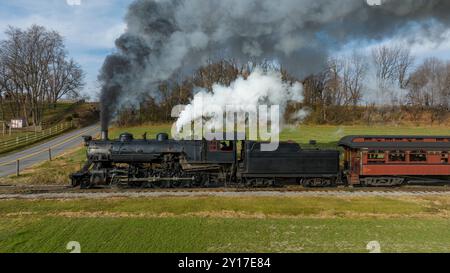 Une locomotive à vapeur classique se déplace le long de voies entourées de champs verdoyants et d'arbres, projetant de la fumée dans le ciel bleu clair par une belle journée. Banque D'Images