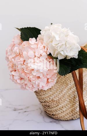 Un panier de paille contient des hortensias roses et blanches avec des feuilles vertes, reposant sur un fond de marbre blanc Banque D'Images