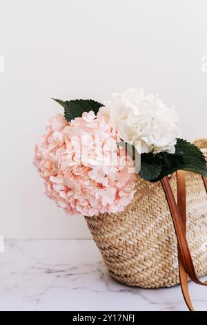 Un panier de paille contient des hortensias roses et blanches avec des feuilles vertes, reposant sur un fond de marbre blanc Banque D'Images